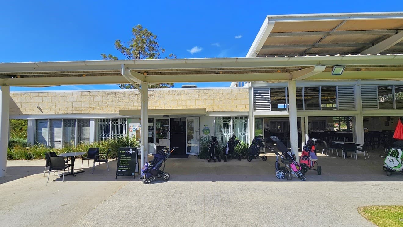 The two-level cream building is a modern structure made of brick and glass. In front, there is a shaded facility housing movable plastic tables and chairs, along with a black freestanding banner. Golf carts are visible. The glass entrance is on the left, featuring the facility banner and stickers. More tables and chairs are arranged inside the building on the right.