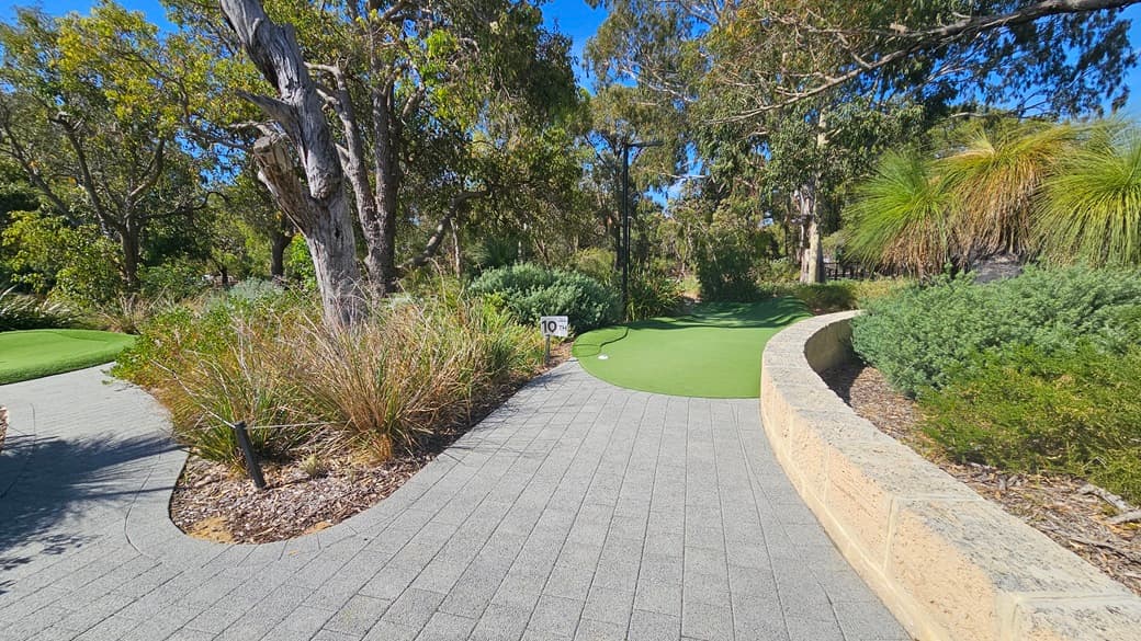 A concrete brick path leading to lower ground. A large, colourful concrete sign is positioned on the right. On the left, a circular brick wall encloses a lamppost. Various types of mature native grass and trees, including Leucophyta shrubs, surround the area.