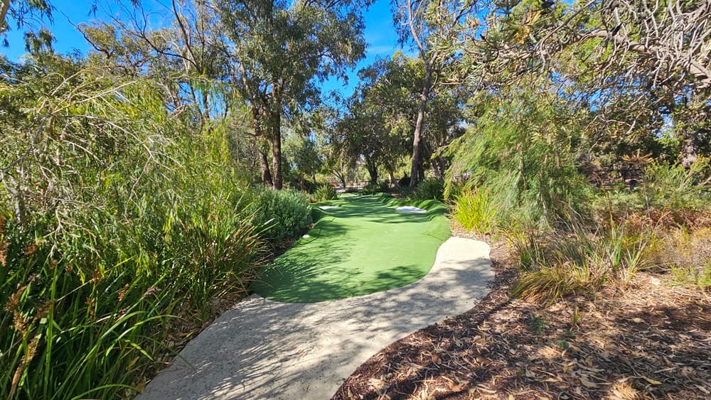 The brick path descends towards the green, surrounded by a landscape of trees, native grass, and shrubs on both sides. To the right, there is a slightly elevated area with soil and native grass.