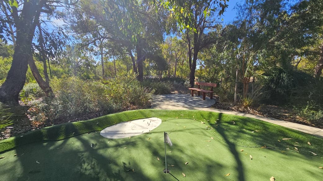 The green is composed of synthetic turf, featuring a bunker in the middle beside a flag. Large trees, native grass, and shrubs encircle the area. A bench is situated beneath a tree in the center. To the right, a descending concrete path is present.
