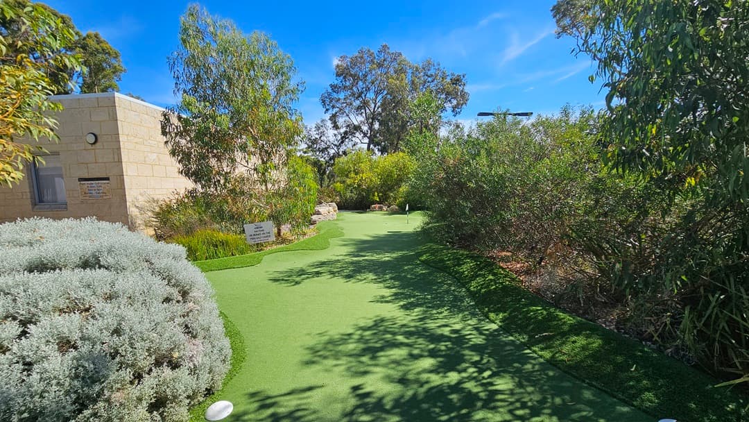 The uneven terrain is paved with concrete bricks and well-kept grass. Ornamental and native plants, along with trees, form a barrier between the grass and concrete. A wooden string fence encloses the area, and a sign reading "1st" is positioned at the center. Tall trees encompass the surroundings.
