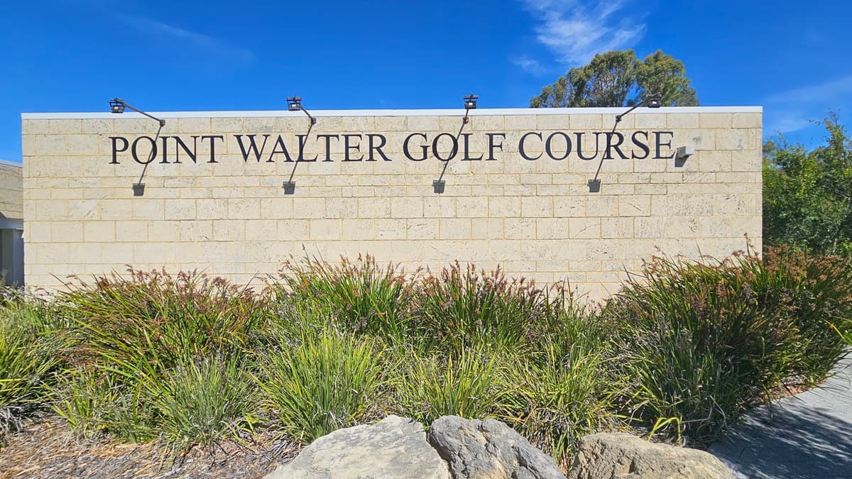A cream brick building displays a prominent sign reading "Point Walter Golf Course" with four attached lights. Below, there's a landscape of native grass and large rocks. On the right, a brick pathway is situated.