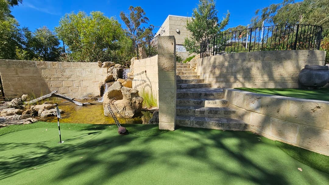 A small waterfall landscape, featuring logs and large rocks, is nestled along a brick wall. To the right are brick stairs leading to a building, surrounded by a metal fence. Well-manicured grass and greenery for the golf hole extend on both the left and right. A banner is positioned on the left, and tall trees shade the area.