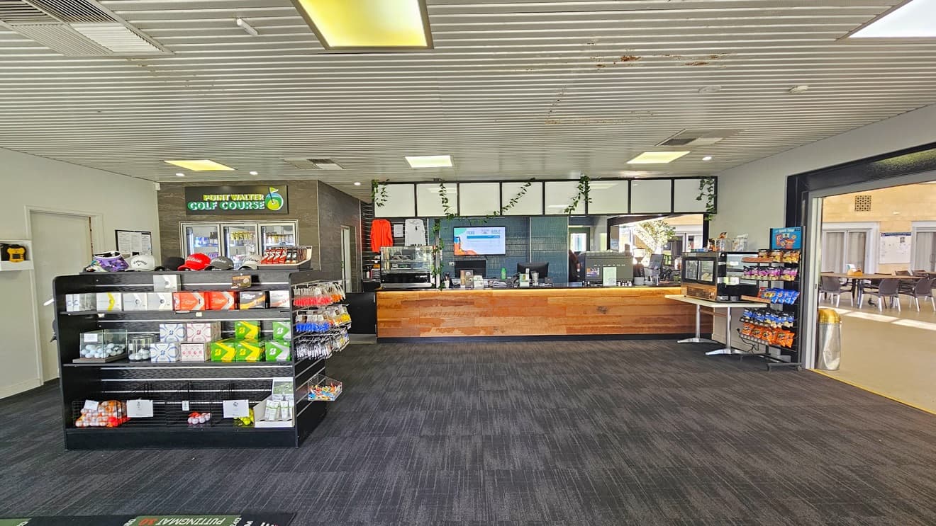 The entrance to the building is through a manual glass door that opens outward. Large banners are affixed to the walls on both sides. A sunscreen dispenser is positioned on the left along with a banner. An anti-slip mat is placed on the doorstep. Native plants are visible on both sides. The ceiling is white with circular fluorescent lights. Inside the facility, the reception area is visible.