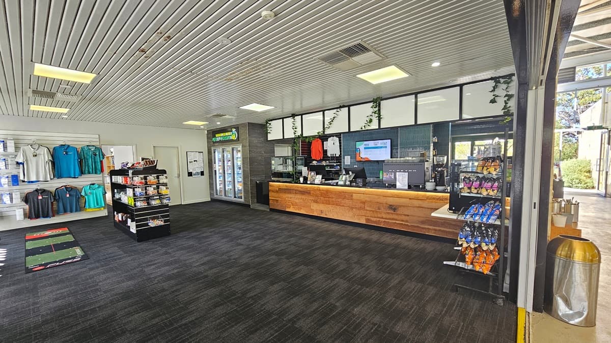 The left side features a display rack with golf items and a large floor mat. The reception desk, with a timber design, showcases retail items and a flat-screen TV. On the counter, a coffee maker and a computer are neatly arranged. Adjacent is a fridge with cold drinks. To the right, a table holds a food rack and a separate one for light snacks. The grey carpeted floor complements the ceiling's corrugated design. Behind a wall, a rubbish bin and a table with kitchen utensils are visible.