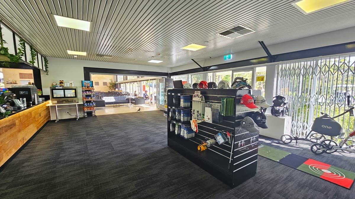 The right side showcases a display rack with golf items and a large floor mat. On the left is the reception desk with a timber design counter. A coffee maker and a computer are neatly arranged on the counter. To the right, a table holds a food rack and a separate one for light snacks. Black leather couches are further in view. The grey carpeted floor complements the ceiling's corrugated design. The right wall is glass with grills.