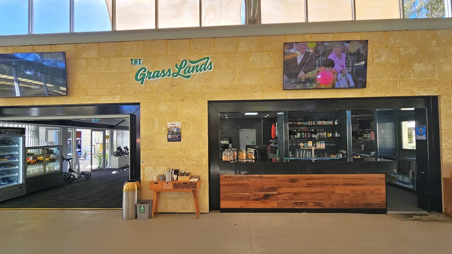To the left is part of the retail area outside the cafe, while the cafe/bar counter, featuring black walls and a timber-designed countertop, is on the right. Inside the bar, bottles of wine and alcohol are displayed. Two large flat-screen TVs are mounted on the cream wall, showcasing the cafe's brand. A table hosting condiments is placed in the middle, with a silver rubbish bin to its left.