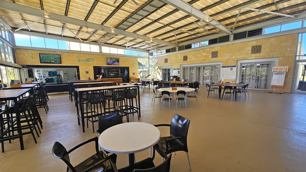 To the left, rows of movable plastic tables and chairs are arranged. On the far left, there's a centre displaying t-shirts for sale. To its right is the cafe and bar counter. Two large flat-screen TVs are mounted on the cream wall. To the right, there are two sets of glass doors. The floor is polished timber, and the ceiling has a sloping industrial design with exposed beams, fluorescent lights, and large ceiling fans.