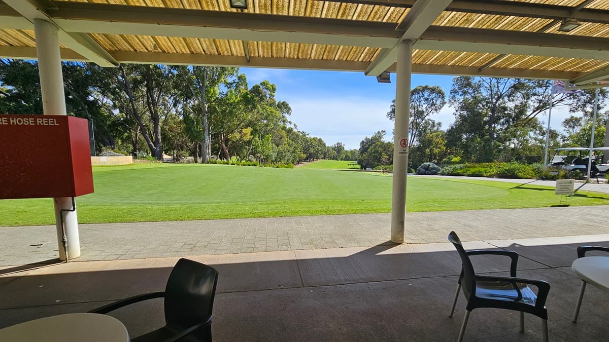 An open space with well-kept green grass. Under the veranda, movable plastic tables and chairs are neatly arranged. The ceiling displays corrugated roofing with exposed metal beams. On the left, a red box houses the fire hose reel. Large trees are visible beyond the grass area.