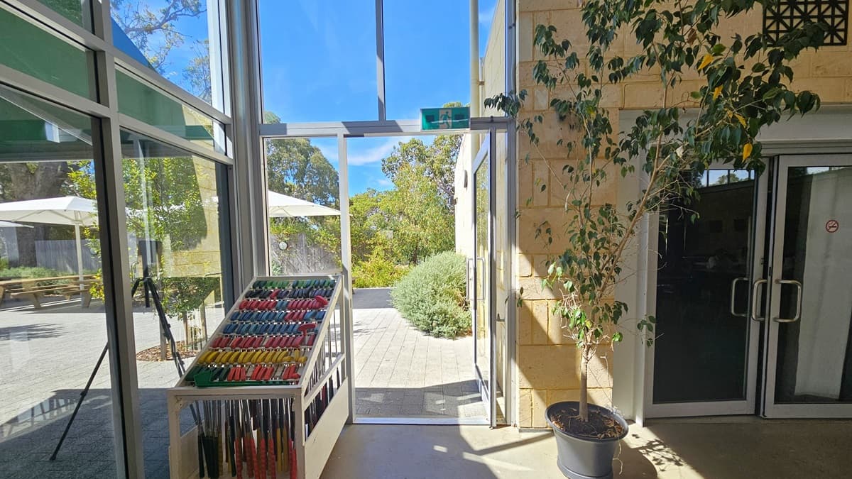 Directly ahead is a glass door which is open and leading to the outside. To the left of this exit are rows of golf clubs lined up by colour and hanging from a metal structure. To the right are glass doors leading to another room. Outside, trees, gardens and umbrellas are visible through the glass windows.