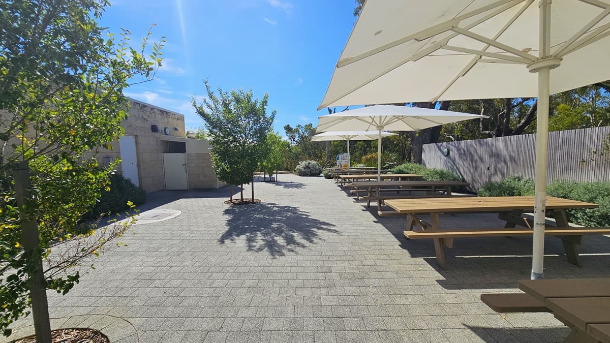 To the left is a low cream building with brick walls. Small trees line the brick pathway. On the right, there's a shaded patio with white umbrella shades and a row of picnic tables. The wall on the right is bordered by plants.