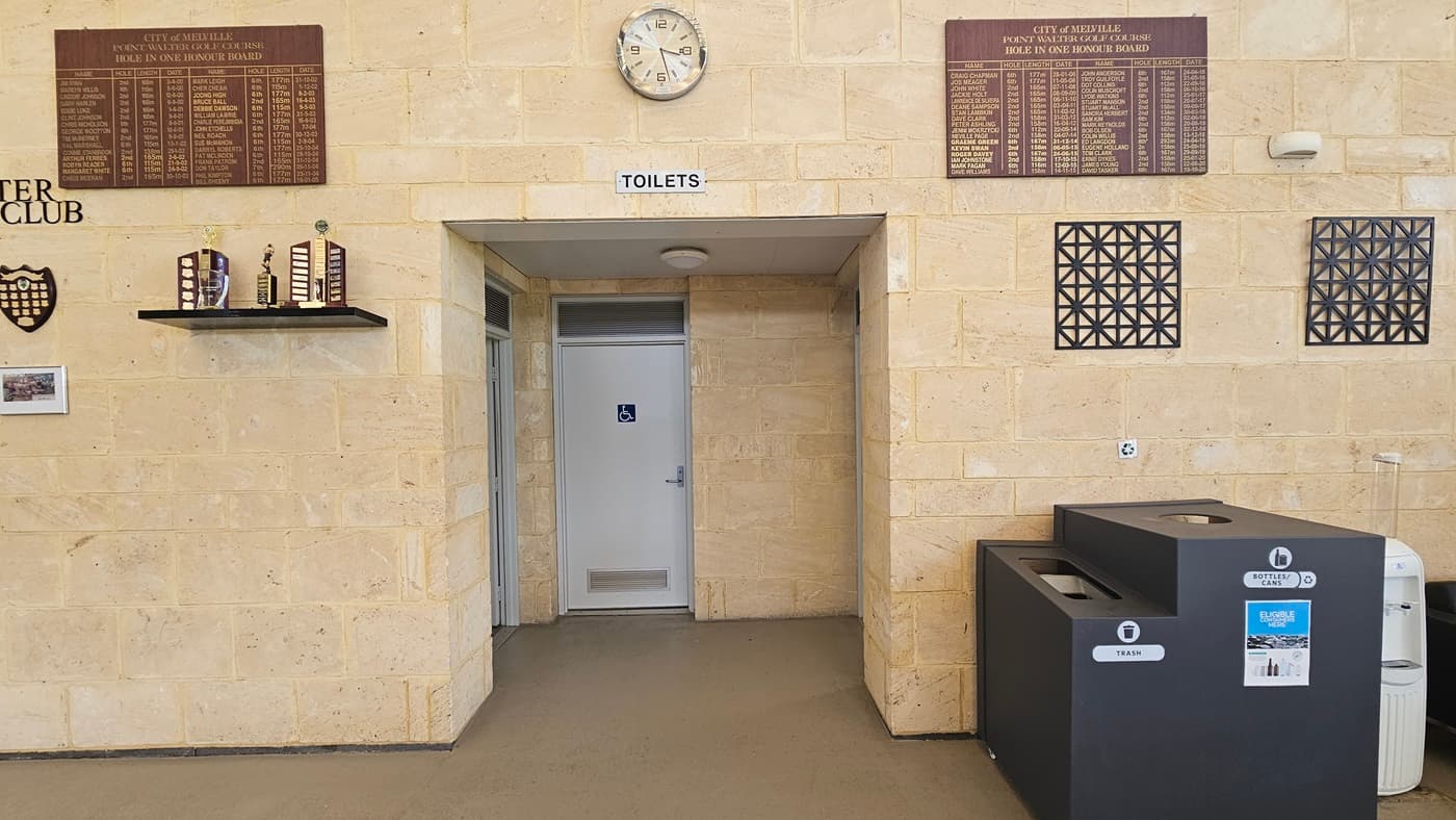 The accessible bathroom, marked with a white door and signage. To the right is a black rubbish bin with a separate hole for recycling. The entire wall is made of cream bricks. Trophies, awards, a brown board featuring "Hole In One Honour Board," a round clock, and wall decorations are displayed.