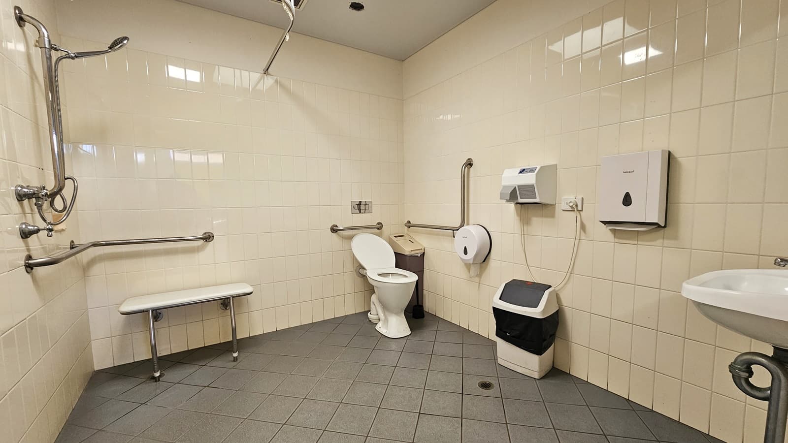The accessible bathroom restroom features a left-installed shower with accompanying handrails. Adjacent to it, a bench chair is placed. In the corner, a toilet bowl and rubbish bin are situated. On the wall, a tissue dispenser, two rubbish bins, and a hand dryer are affixed, alongside a wash basin. The wall exhibits white tiles, and the ceiling and floor are adorned with white and grey tiles, respectively.