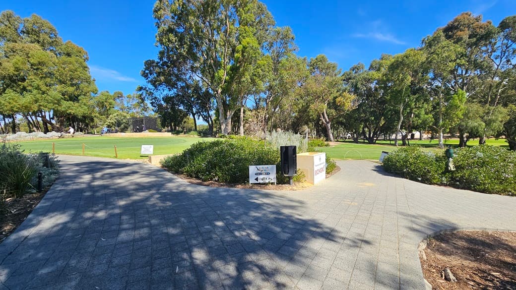 The concrete brick pathway divides; both lead to a vast green and grassy area with tall trees and shrubs. In the middle is a mini landscape with directional signages, a golf hole sponsor, and a garden lamp. The path on the left elevates towards the entrance of the building. A wooden post stands on the right along with a banner. The path on the right descends to the golf area.