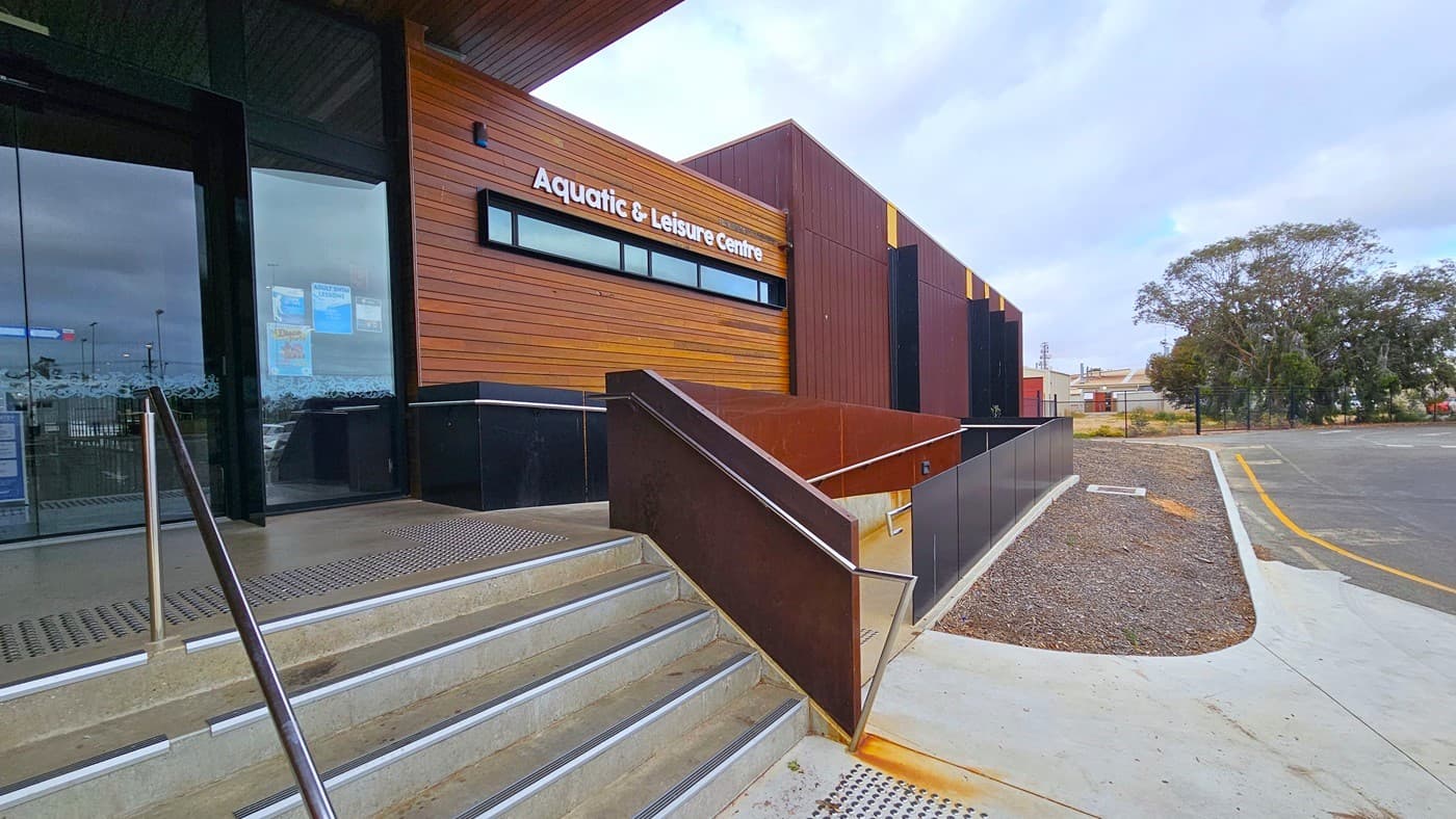 In the centre stands a structure with modern architecture, showcasing a sloping yellow roofline, timber walls, and a warm brown and yellow timber ceiling. Prominently displayed at the front is the facility signage. Two sets of steps with handrails, each equipped with truncated domes on the floor, are located in the middle. On the right, there is a ramp entry with a railing. An empty concrete planter box and a section of the parking area are visible on the right, along with mature trees.