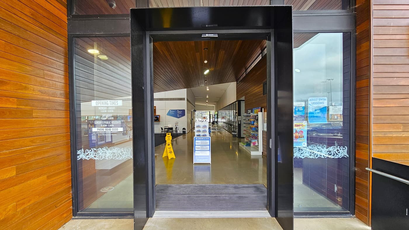 The entrance features an automatic sliding glass door with stickers displaying store hours and posters affixed to it. A non-slippery mat is placed at the entrance. Both walls have a timber design that extends inside the building, including the ceiling. Inside, a freestanding sign is positioned in the center, along with a yellow safety sign and a retail rack filled with swimming accessories is placed on the right.