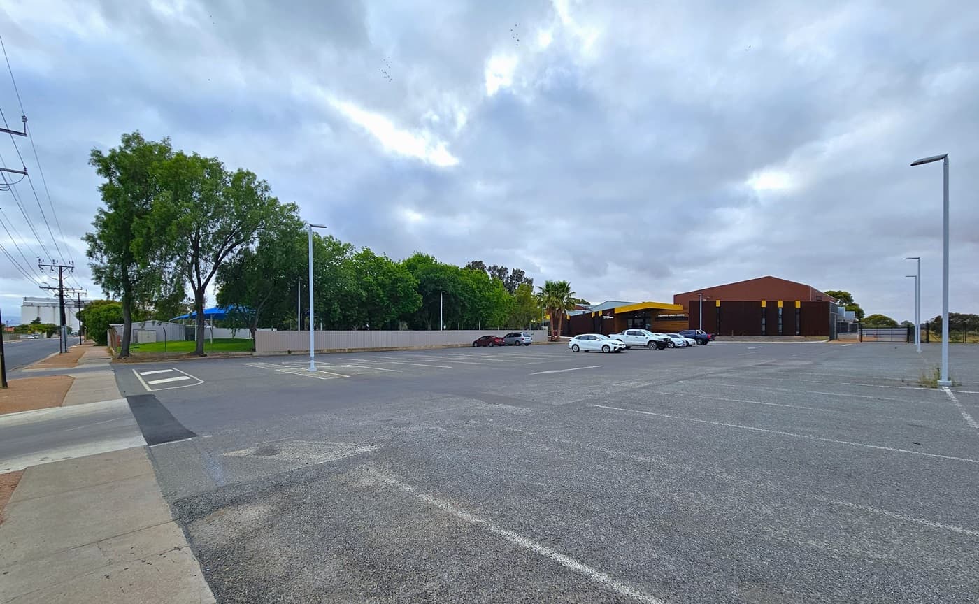 This is the expansive concrete car park adjacent to the Port Pirie Aquatic and Leisure Centre facility. The parking area is marked with white lines, and tall light posts are strategically positioned. In the middle, a few cars are parked. On the left is the main road, and further ahead, tall trees border the road and the grassy area.