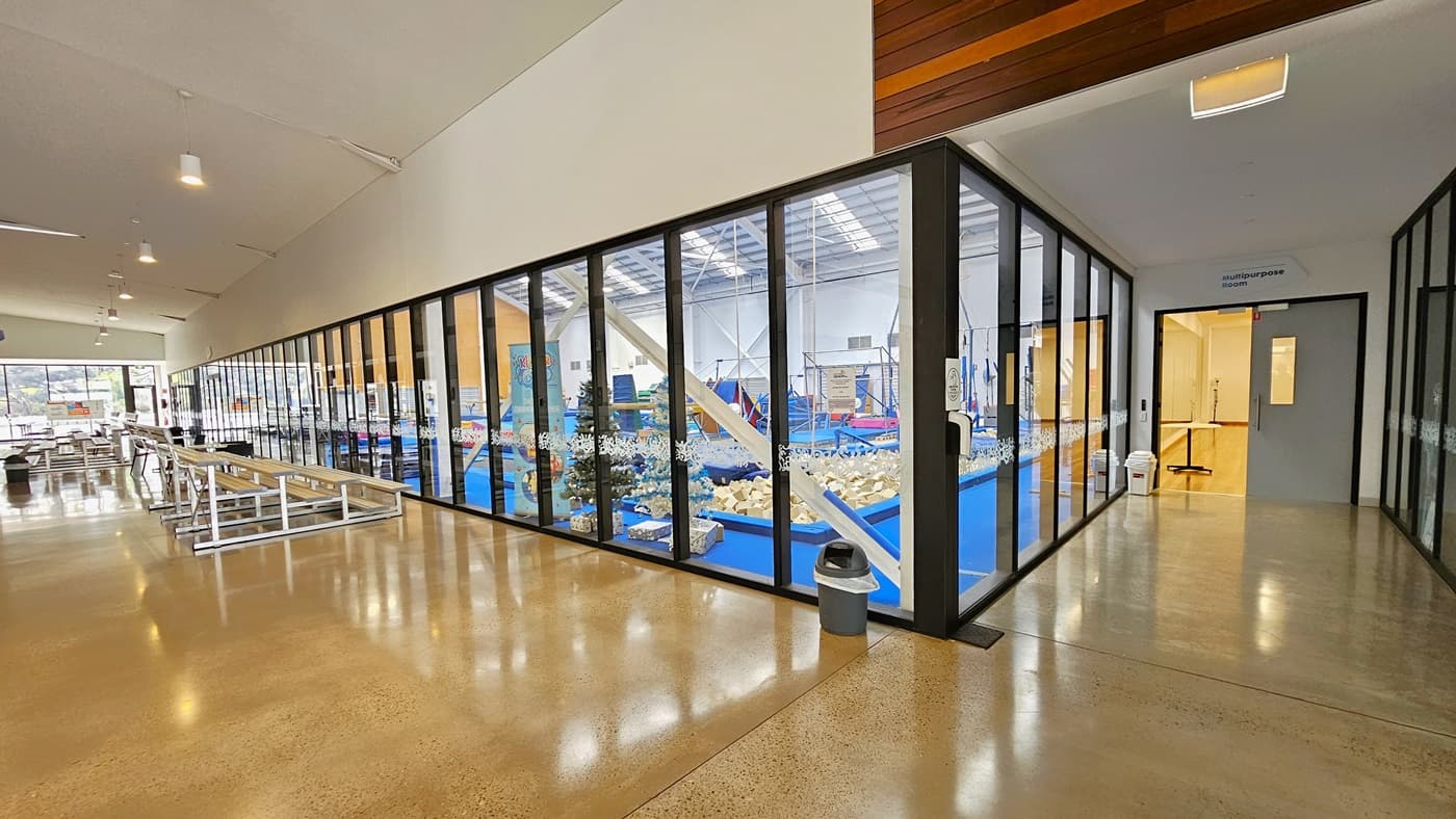 To the left is a corridor that leads to the aquatic facilities. Aluminum-tiered seatings are positioned in front of the gymnastics hall. To the right is another corridor leading to the multi-purpose hall. The floor is made of polished concrete, and the ceiling is all white with light fixtures hanging. Rubbish bins are placed in the corners.
