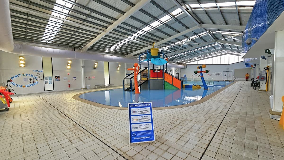The entrance to the indoor Aquatic facility is through automatic glass sliding doors with posters. To the left, there's a poster and a white rubbish bin. Inside, to the right is the splash zone area with colorful slides and water features. To the left is a floor-to-ceiling glass wall with a view of the outdoor pool. A few free-standing banners are present, and the ceiling features exposed metal beams, insulated foams, and natural skylights.