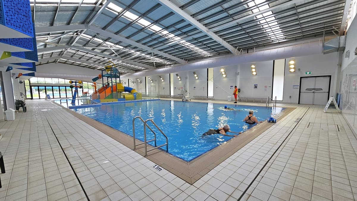 The pool is equipped with stainless steel hand ramps on the left and right sides. On the right side of the pool are the accessible bathrooms. The walls on the left side of the pool have a vibrant blue lining. Plastic chairs are placed on the pool deck against the wall along with a rubbish bin. There are a few free-standing banners in the corner. The ceiling has exposed metal beams, insulated foams, and natural skylights. Further is the Splash Pool featuring a colourful slide.