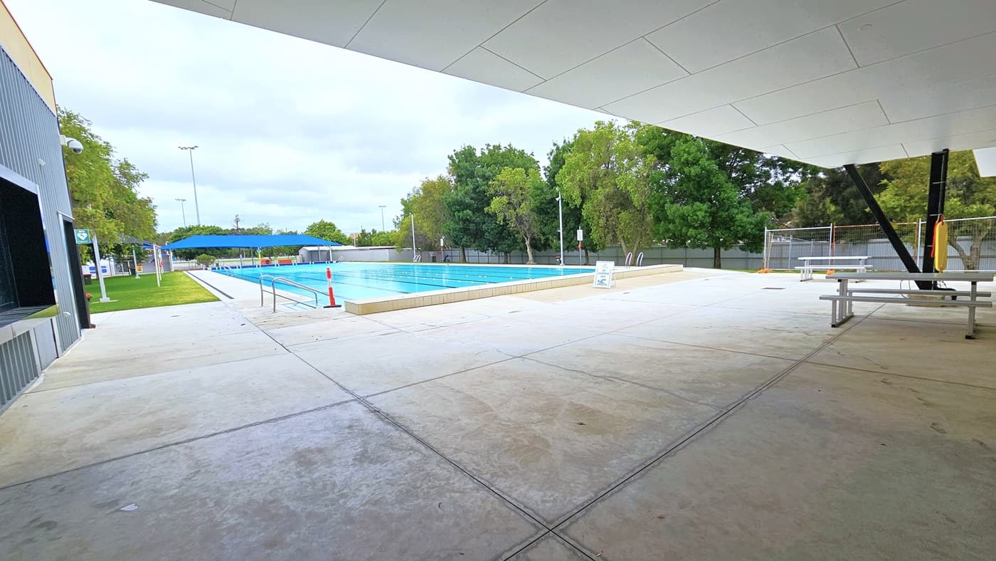 The outdoor Aquatic Centre entrance features an automatic glass sliding door with a grey non-slip mat. A Fire Exit sign is affixed on top and white rubbish bin is positioned on the right. The 7-lane pool area is on the left, and a shaded seating area with bench chairs is on the right, near the glass wall. The area is enclosed by a fence and surrounded by trees.