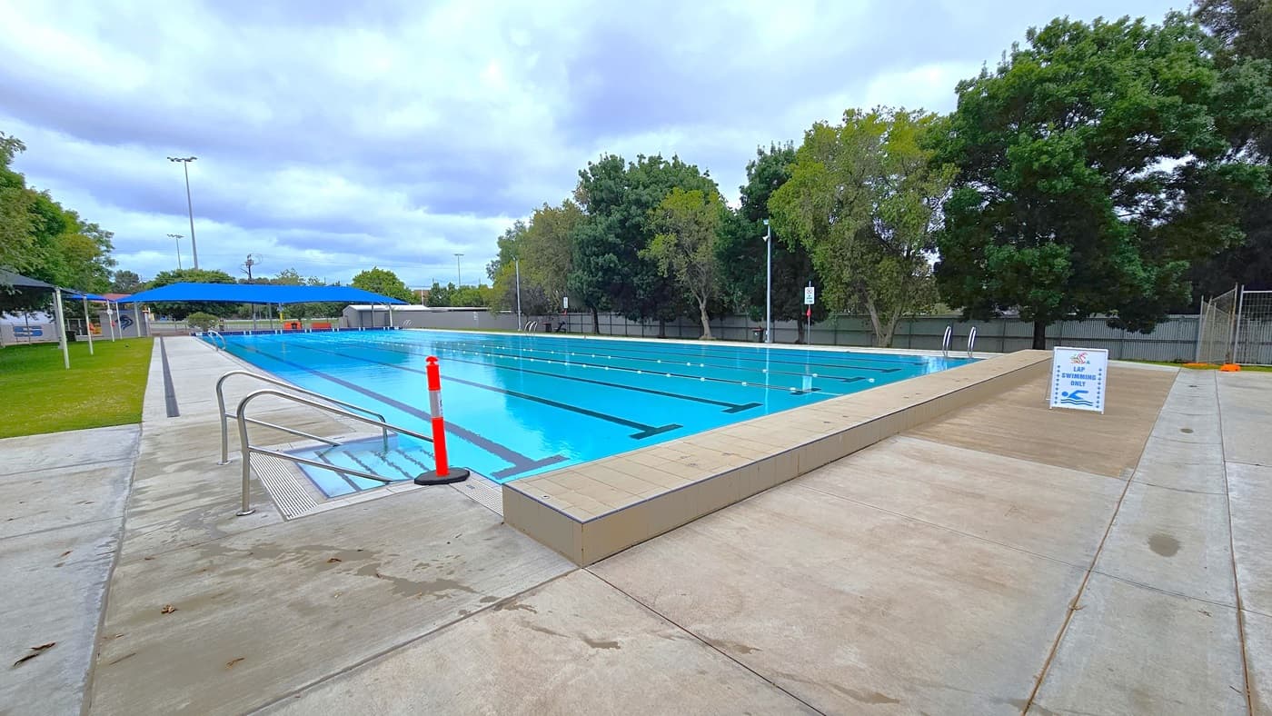 In the middle is the 7-lane, 50-metre swimming pool equipped with handrails on each corner. An orange bollard is positioned at the corner. The pool coping is made of cream tiles, and a freestanding banner stands in the middle of the pool deck. On the left is a grassy area with shade cloths that extend towards the other side of the pool. On the right, there are chain fences and corrugated walls surrounding the area. Tall trees border the right side of the pool.