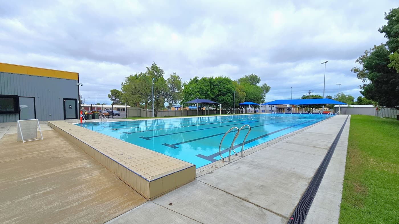 In the middle is the 7-lane, 50-metre swimming pool equipped with handrails on each corner. An orange bollard is positioned at the corner. The pool coping is made of cream tiles, and a freestanding banner stands in the middle of the pool deck. On the left is a grassy area with shade cloths that extend towards the other side of the pool. On the right, there are chain fences and corrugated walls surrounding the area. Tall trees border the right side of the pool.