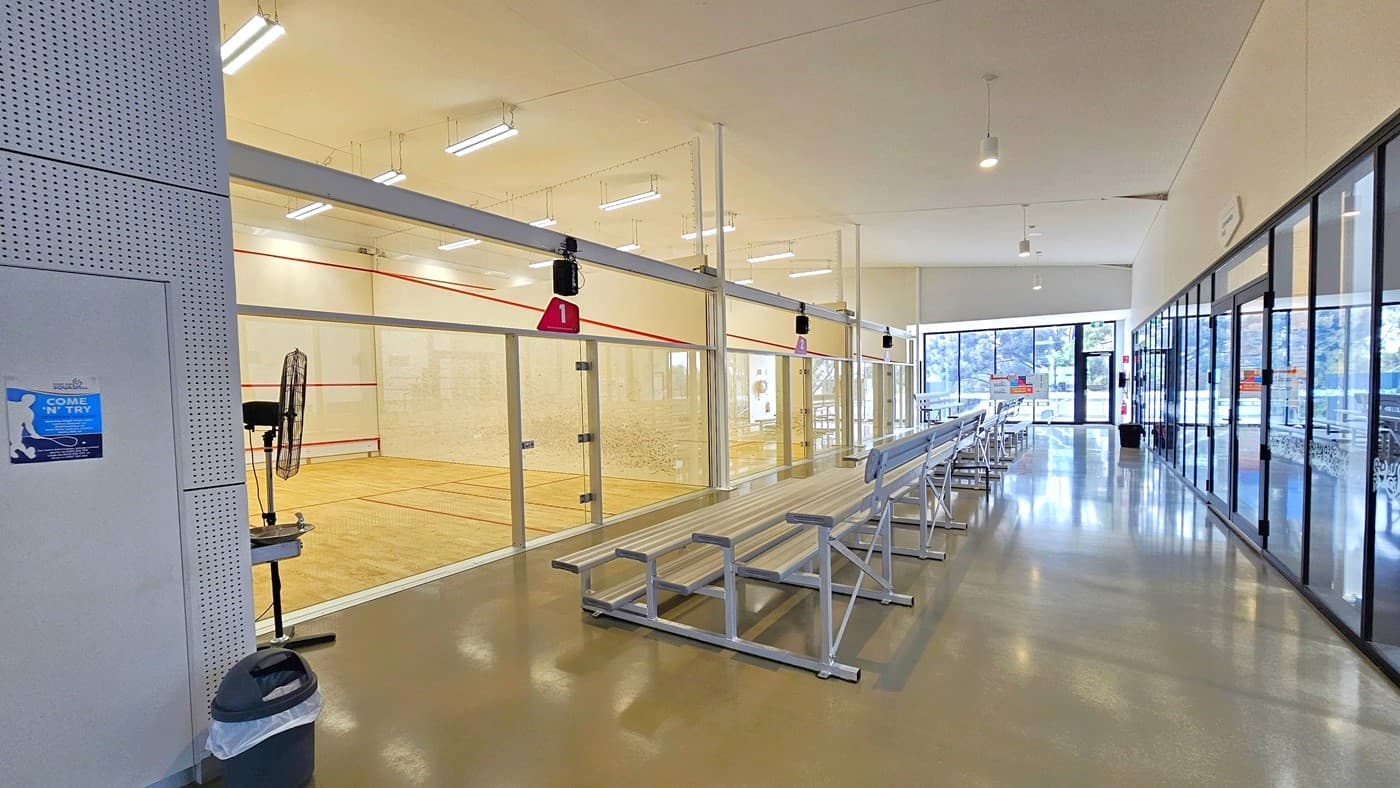 In the lobby's middle, tiered seating provides a view of the squash courts. A stand fan sits in the left corner beside a black rubbish bin. The squash court boasts timber floors, white walls, and a ceiling with fluorescent lights. More tiered seating lines the lobby, and on the right, the gymnasium area is enclosed by a glass wall extending towards the lobby's end.