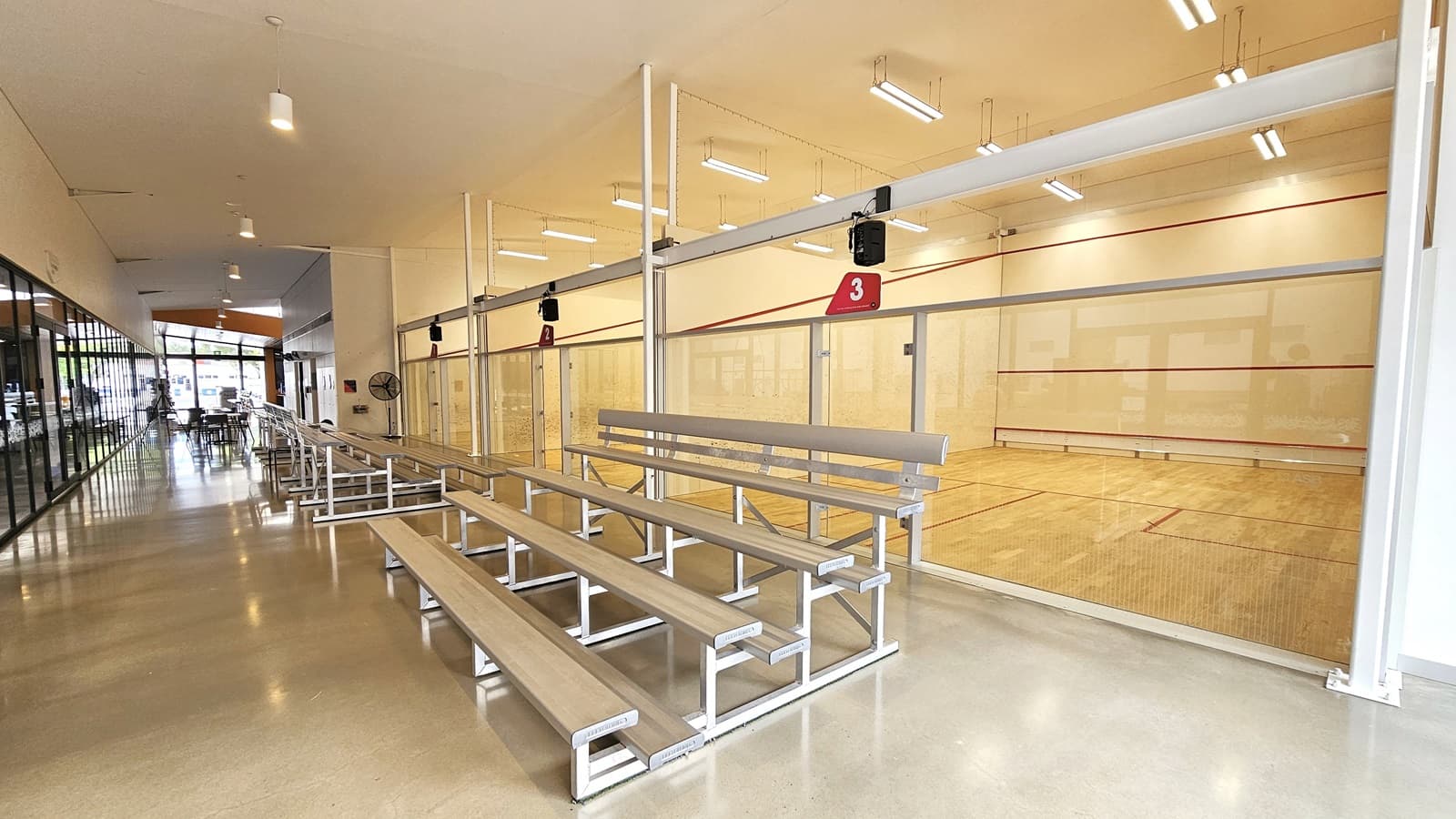 To the right, three squash courts are positioned: Court 1 on the farthest, Court 2 in the middle, and Court 3 on the right. Each court shares identical features—timber floors with vibrant red markings, white ceilings adorned with double fluorescent lights, and white walls. Each court has its own glass door entrance. Tiered aluminum seating faces the court and the gymnastics hall on the left. The glass wall on the left extends towards the reception lobby.
