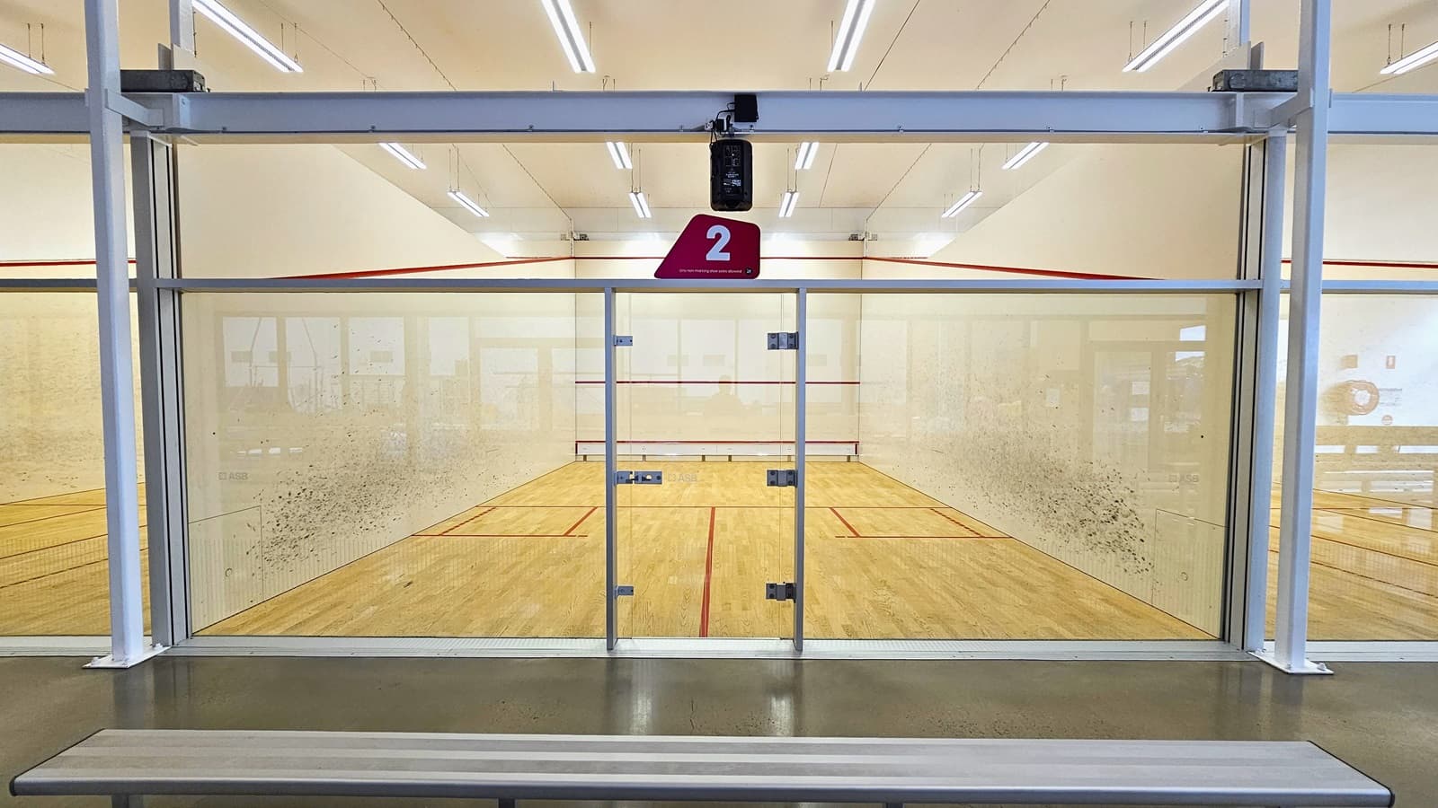 A detailed look at Squash Court #2 reveals an inward-opening glass door positioned in the middle for access. The court features timber floors with vibrant red markings for the game. An additional red mark lines the upper wall surrounding the space. Black markings adorn both sides of the wall, and the ceiling is white with double fluorescent lights. To the left is Court #1, and to the right is Court #3. Tiered aluminum seating is positioned in front.