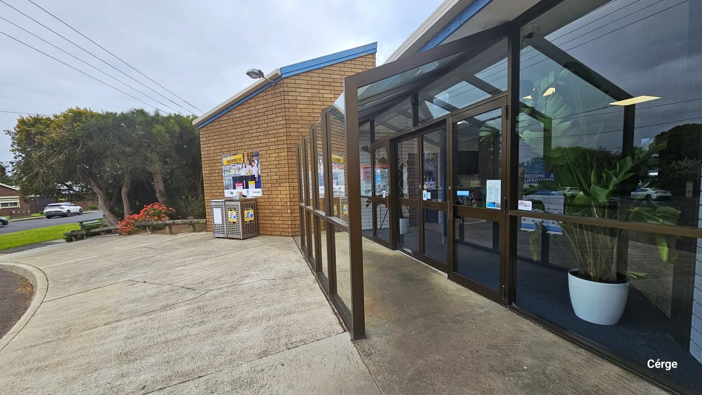 A closer look at the glass entrance doors with chocolate steel frames. Inside are two white potted plants that have banana-like leaves, a grey floor, and a white brick wall. Mounted on top of the left brown brick wall outside, right above the RECYCLE IT poster and perforated bins, is an outdoor lighting fixture. The facility has a garden with red flowers and tall trees with wide canopy cover.