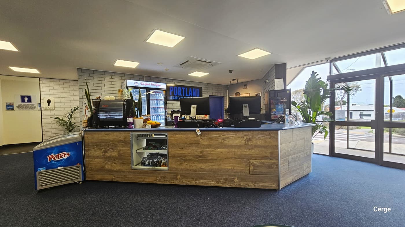 Reception desk, accessible change room, and available seating after passing through the glass entrance doors of Portland Leisure Centre. The facility has a grey natural stone floor, white brick walls, and a white ceiling with square ceiling fluorescents that emit warm light. On the right is the wooden reception desk with a light blue countertop. A blue banner in the center reads “Become a Lifeguard” with a back image of a lifeguard wearing a red and yellow uniform.