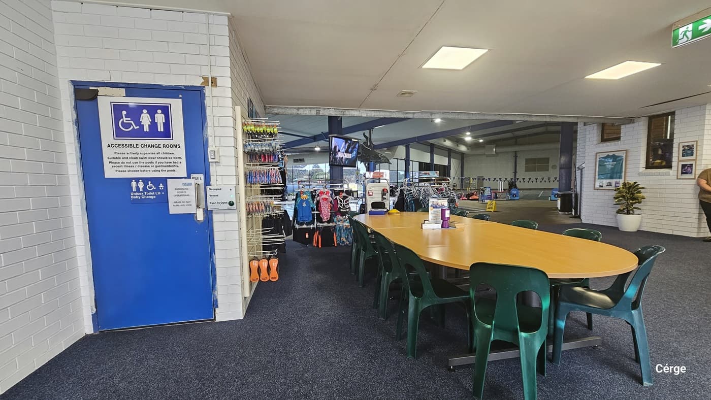 Available seating near the Accessible Change Rooms and retail offering of swimwears and swimming accessories. The seating area has green, plastic, monobloc chairs around a wooden elongated table. On the left white brick wall is a blue door with an “Accessible Change Rooms” grey sign and a Unisex Toilet LH + Baby Change blue sign. The door can be operated through a grey metal plate that contains the green button for opening the door and two lights for occupied and vacant.