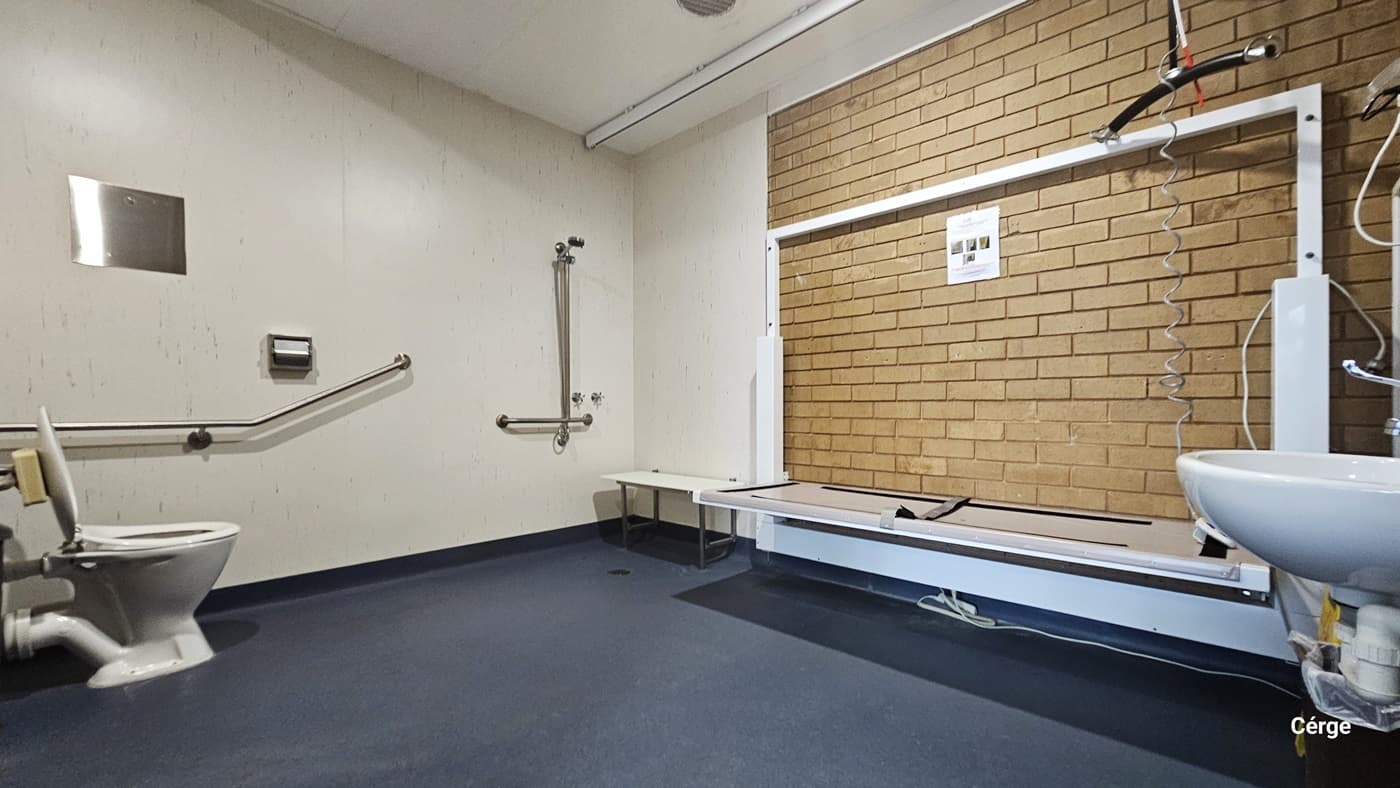 Inside the accessible change room at Portland Recreation Centre. The room has a grey natural stone floor, a white ceiling, and a brown brick on the right, while the other walls are white. On the left end is the toilet with metal grab bars. On the right corner is the handheld shower head, a white shower seat, and an adult change table. Near the door, to the right, is a wash basin.