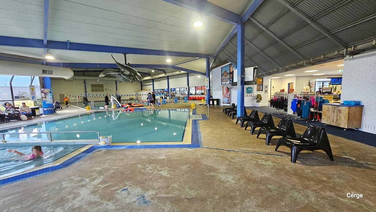 Multipurpose indoor pool at Portland Leisure and Aquatic Centre. The first pools accessible after the reception are separated by a white elevated section with stainless steel railings in the middle. Both pools have a cream and blue pool coping. Grey whales and sharks hang from the ceiling. On the right pool deck are black plastic chairs perfectly aligned a few meters away from each other. To the back is a white pool hoist.