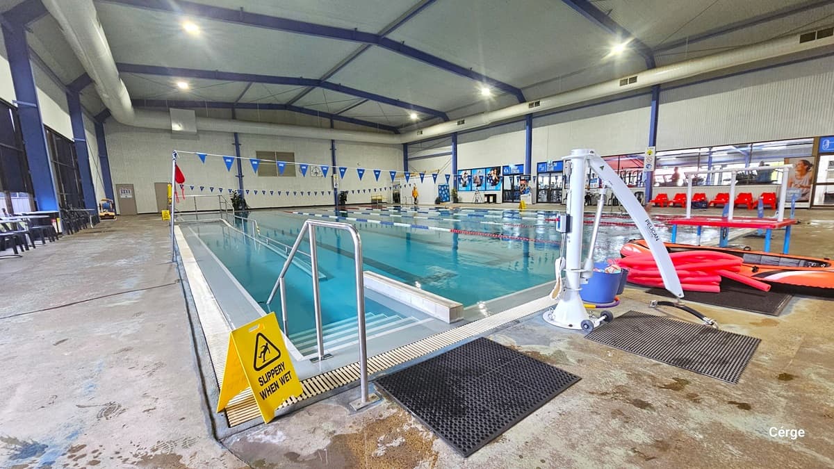 Pool stairs, ramp access, and pool hoist at Portland Leisure and Aquatic Centre. Before entering the pool stairs, there is a black rubber mat and a yellow SLIPPERY WHEN WET floor sign. On the right are the white pool hoist, red pool noodles, and an orange floating device. On the bottom pool floor is the bold blue center T line that helps the swimmer stay at the center. To the back wall, on the left side, are two brown doors.