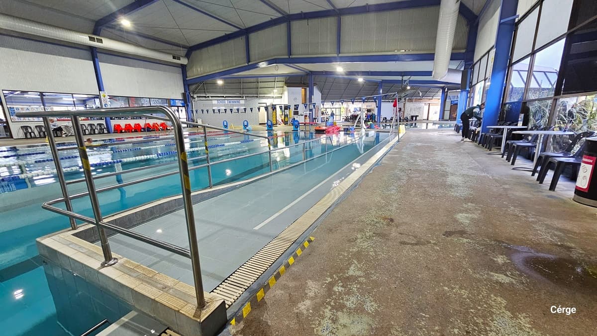 Ramp access with metal handrails in the 25-meter, 5-lane indoor pool. On the right pool deck is a silver circular metal table and black monobloc plastic chairs with an outdoor nature view through the glass windows. Each lane is separated by the white, red, and blue floating rope lanes. On the bottom pool floor is the bold blue center T line that helps the swimmer stay at the center. On the left pool deck are black and red plastic chairs.