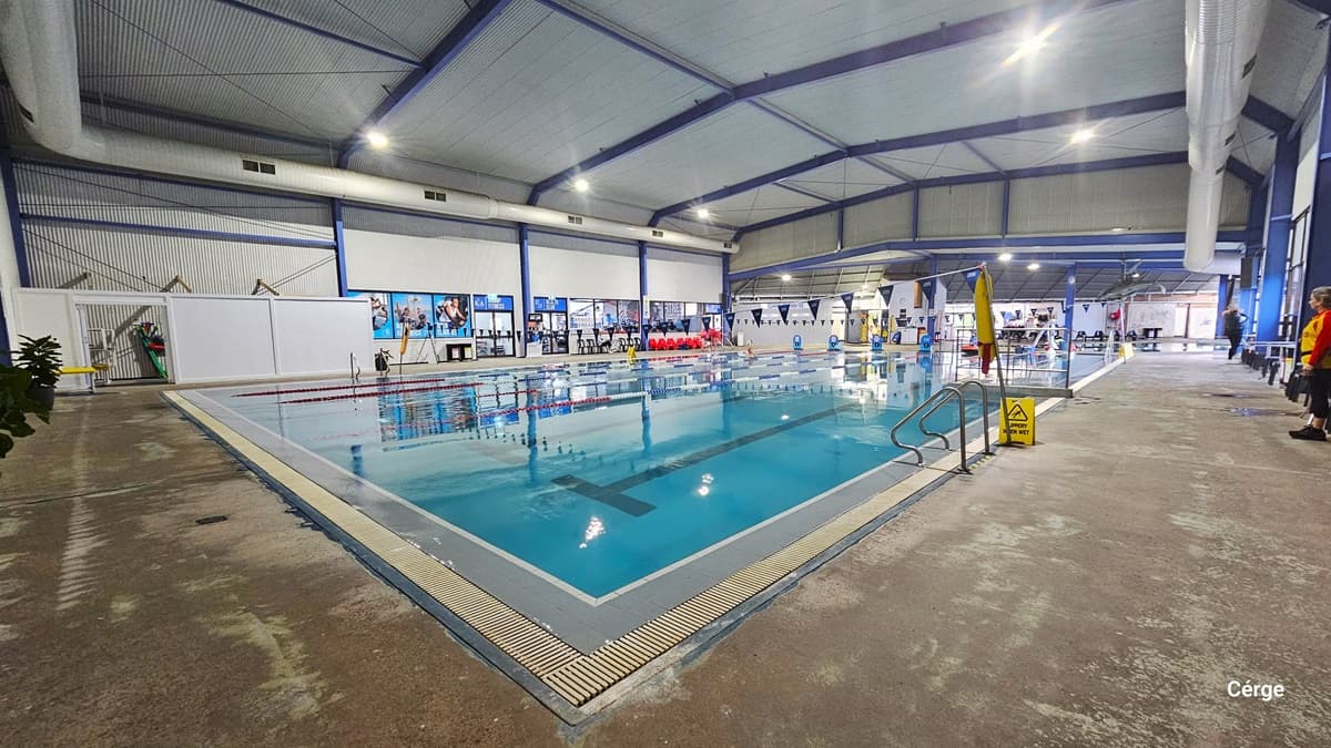 25m, 5-lane swimming pool with ramp access and pool hoist in Portland Leisure and Aquatic Centre. On the right pool coping are two curved stainless steel grab rails near a yellow SLIPPERY WHEN WET sign. The pool lanes are separated by red, white, and blue floating rope lanes. Intersecting the pool are strings of triangular blue flags hanging above. The facility is a large steel structure with a white corrugated iron ceiling, blue steel beams, and large white ventilation ducts on both corners.