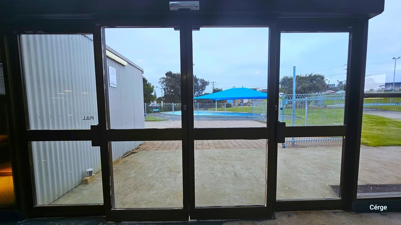 Automatic glass sliding doors to the Outdoor Aquatic Centre at Portland Leisure and Aquatic Centre. The glass doors have dark metal frames. Seen through the glass doors is the grey corrugated wall to the left, a concrete outdoor floor, a brown brick floor, a sky blue fence, and a blue shade sail over the outdoor pool. The grass is well-maintained next to the fence.