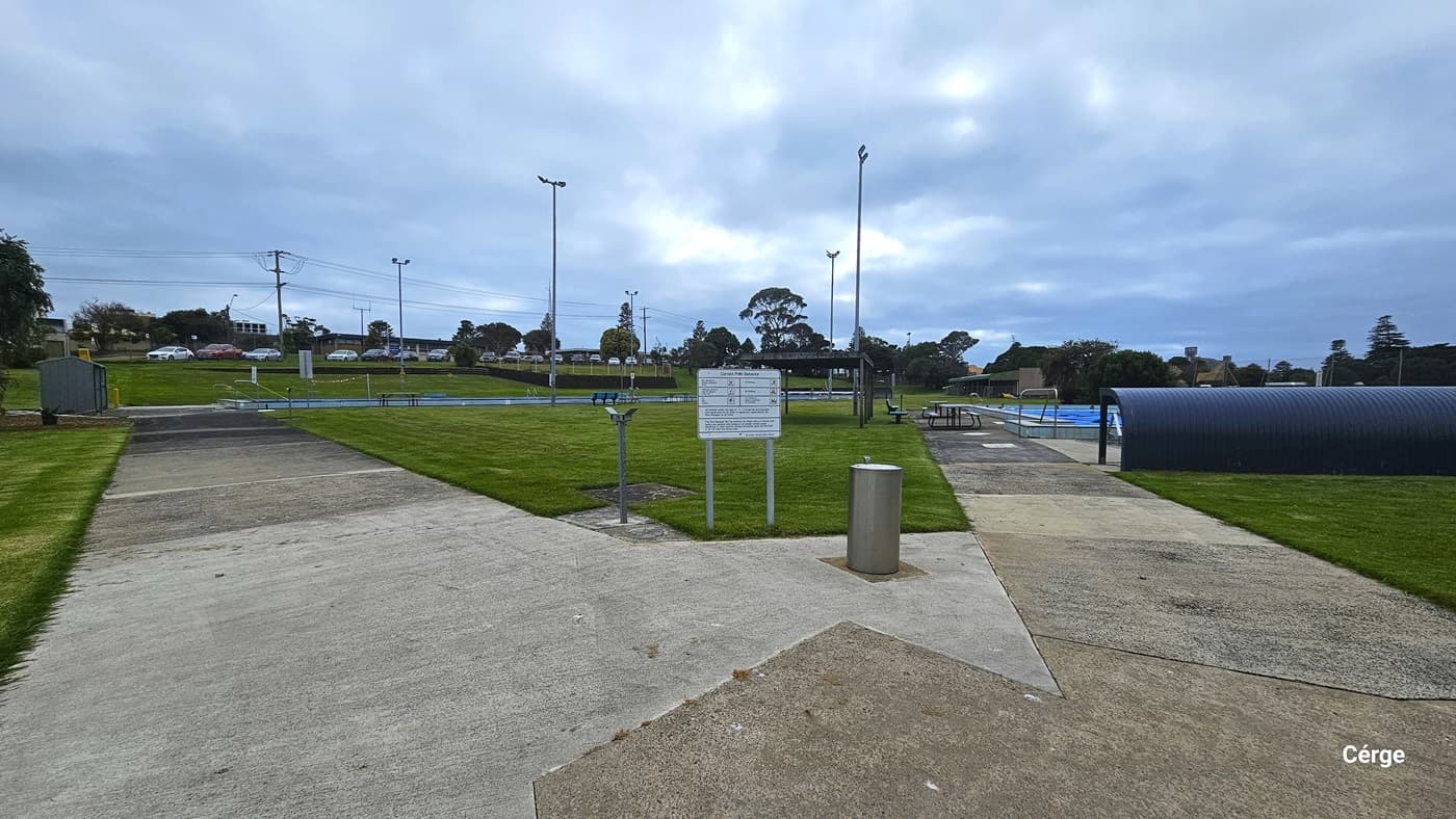 Concrete pathway leading to the 25m and 50m swimming pools heated to 28 degrees. In the middle greenery that forms a triangular grass section is a white sign near a stainless steel garbage bin. On the right side is a grey corrugated curvature that offers shade from the direct heat of the sun. Picnic tables, benches, and a waiting area with a roof can be seen on the left pool deck.