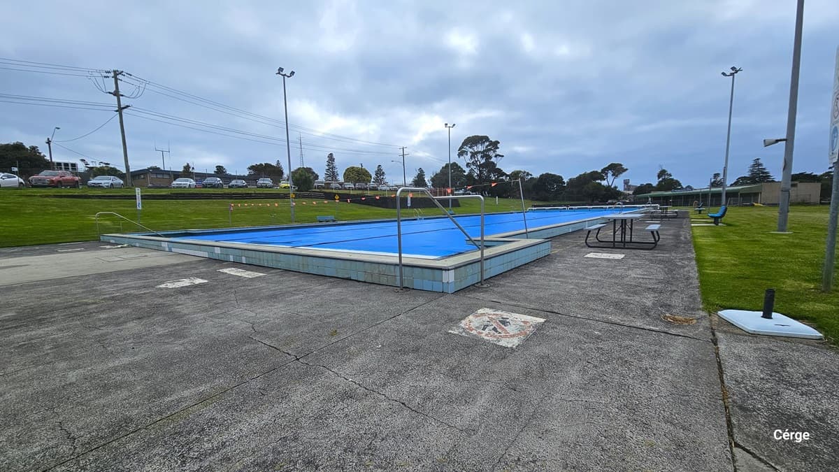 The 25m swimming pool at Portland Leisure and Aquatic Centre. The pool coping is a combination of white tile with a blue accent. A string of orange triangular flags are hanging perpendicular to the pool. Picnic-style tables and chairs are on the right pool deck. Tall lamp posts are available on all sides of the area. Seen from this position is the parking lot filled with varying colors of cars.