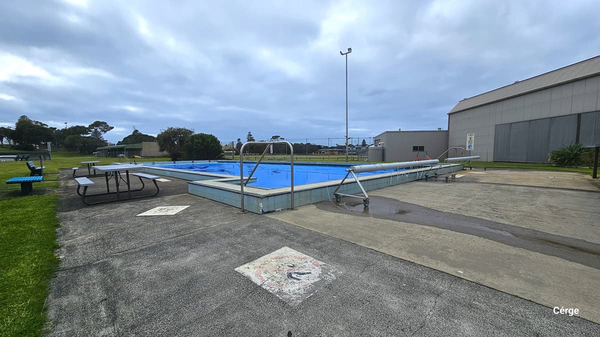 The 50m swimming pool at Portland Leisure and Aquatic Centre. On the left pool deck are light blue picnic-style tables and sky blue benches. On one end of the pool are two rolls of plastics wrapped around a movable metal frame with black wheels. The facility has light brown and grey corrugated walls. The outdoor pools are surrounded by lush grass with an overlooking view of the residential area.