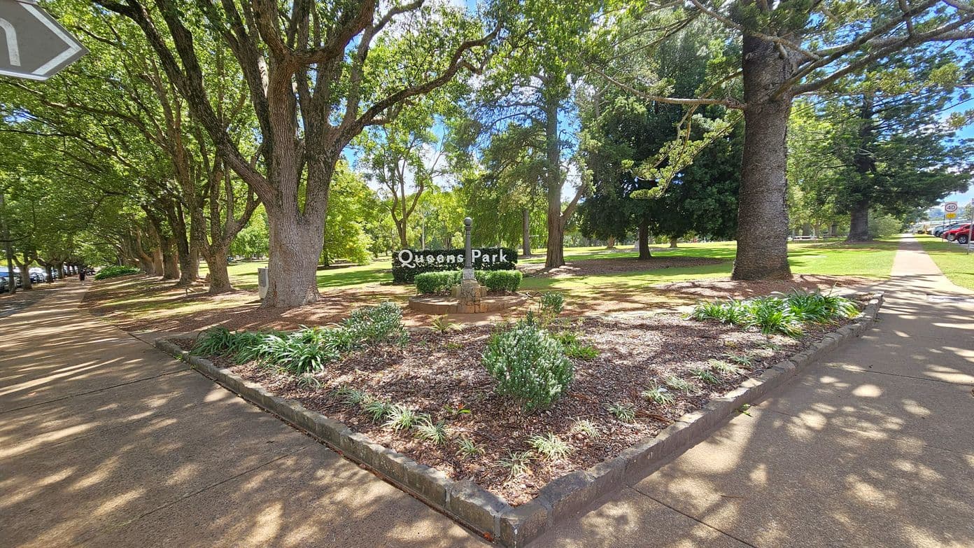 A park scene with a central garden bed featuring a stone structure and a sign reading "Queens Park". Tall trees with dense foliage line both sides, casting shadows on the ground. The left side has a paved walkway bordered by trees, while the right side shows another path with parked cars visible in the distance. The ground is covered with mulch and small plants, and the area is well-maintained.