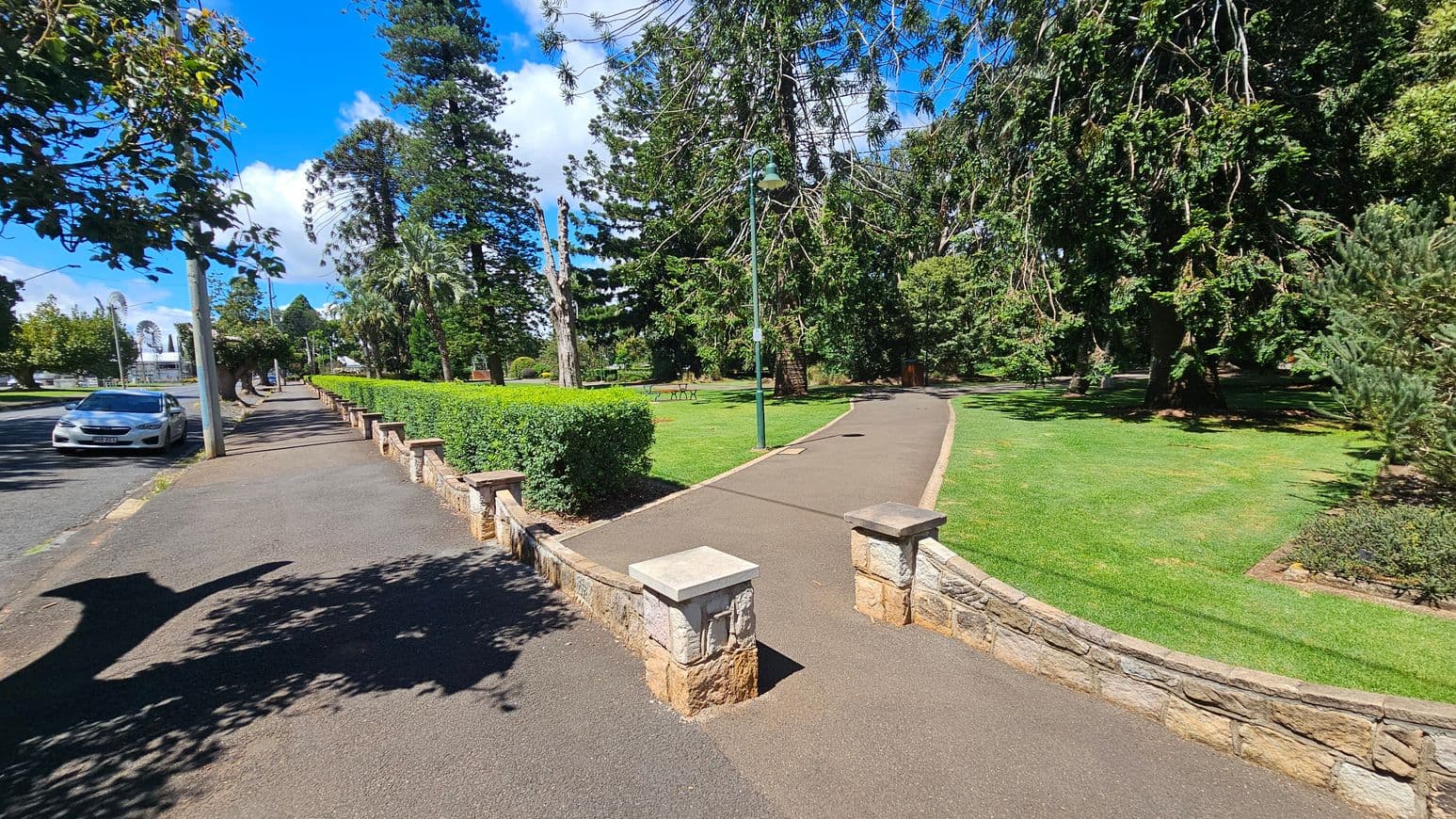 A paved path curves through a park with lush green grass and tall trees on the right. A stone wall with pillars borders the path. On the left, a pavement runs alongside a road where a white car is parked. A hedge separates the pavement from the park. Street lamps line both the path and the pavement. Shadows of trees are cast on the pavement, and the sky is clear and blue.