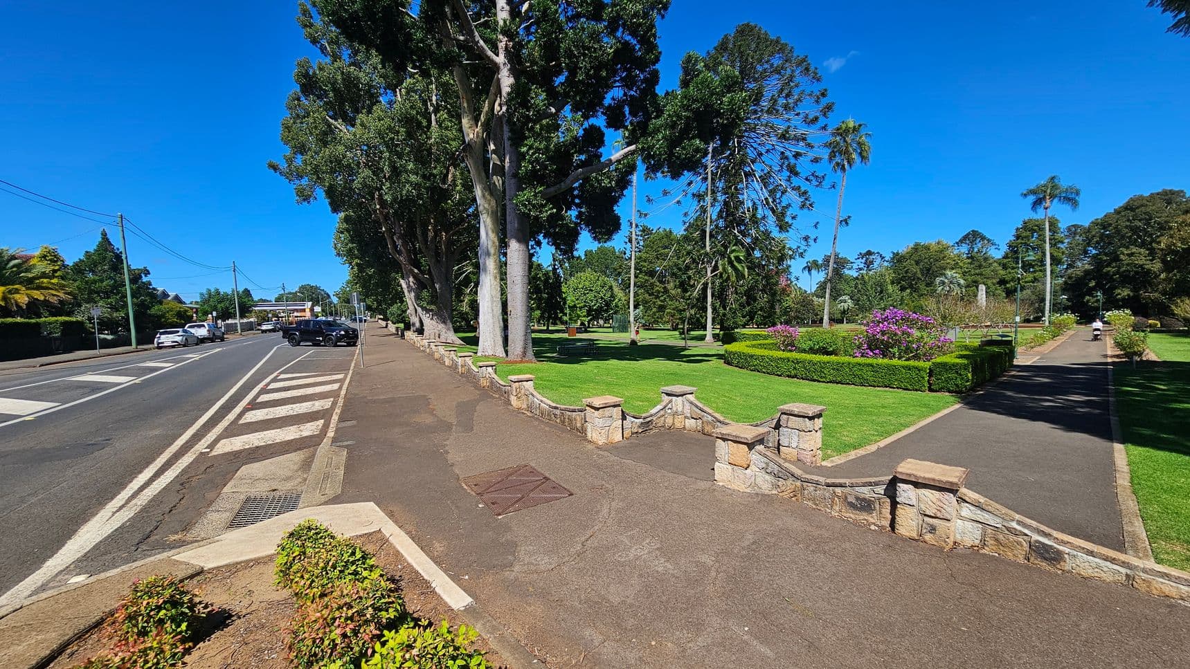 A paved road runs along the left side with parked cars and a pedestrian crossing. On the right, a park features a stone fence, large trees, manicured lawns, and flowerbeds with purple blooms. A path runs through the park, where a person is walking in the distance. The foreground has a small patch of shrubs, and the background is filled with more trees and greenery.
