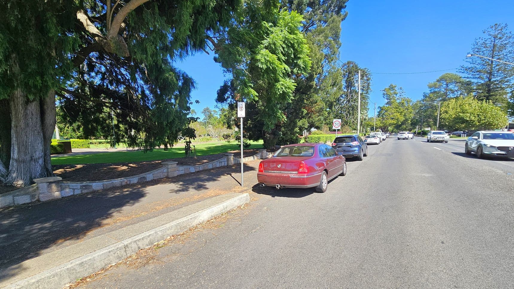 A street scene with a red car parked on the left side near a pavement. A large tree with dense foliage provides shade over the pavement and part of the road. A "No Parking" sign is visible next to the car. Further down the street, several other cars are parked along both sides. The road is lined with trees and a speed limit sign indicating 50 km/h. The pavement is bordered by a low stone wall, and the area appears to be residential or park-like.