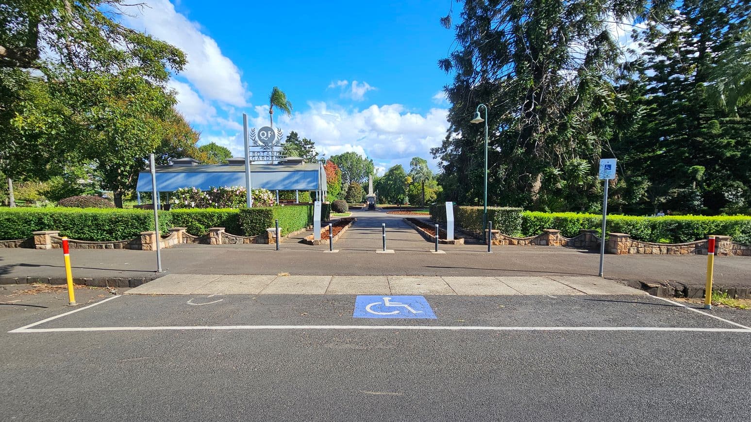 A pathway leads into a garden area, flanked by neatly trimmed hedges and tall trees on both sides. In the foreground, there is a marked disabled parking space on the asphalt. A sign with the letters "QF" is visible on the left, near a small building with a grey roof. The background features more greenery and a clear blue sky. A lamp post stands on the right side of the path.