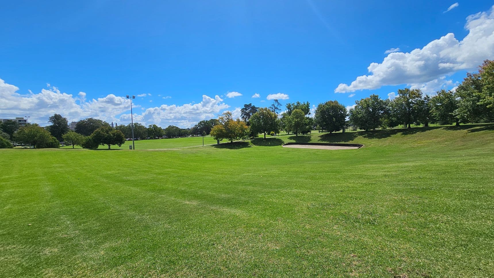 A wide expanse of green grass covers the foreground, leading to a gentle slope. In the background, a row of trees lines the horizon, with a few scattered trees in the middle ground. A light pole stands to the left, and a small structure or bench is visible near the trees. The sky is clear with some clouds.
