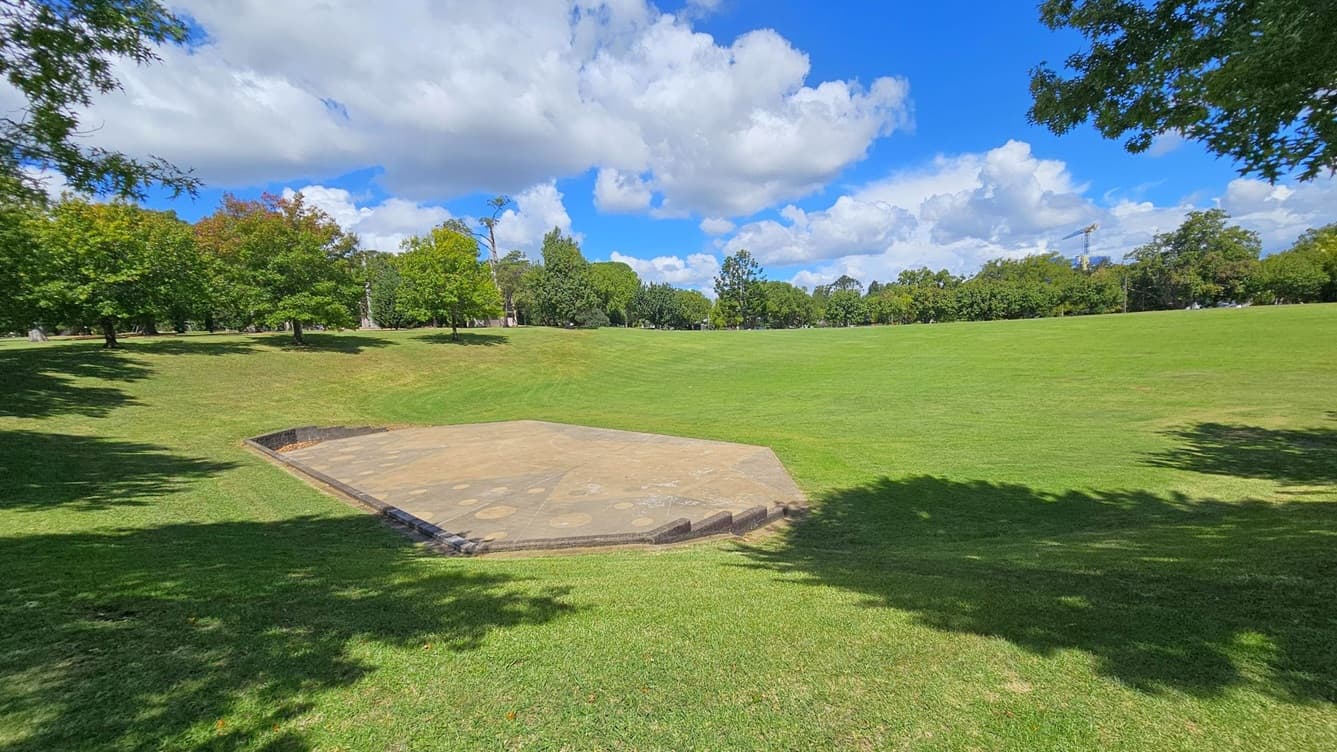 A grassy park landscape with a hexagonal concrete platform in the foreground. The platform is surrounded by well-maintained grass. In the background, there are numerous trees with green and some autumnal leaves. Shadows of trees are cast on the grass, and a construction crane is visible in the distance on the right. The sky is partly cloudy.