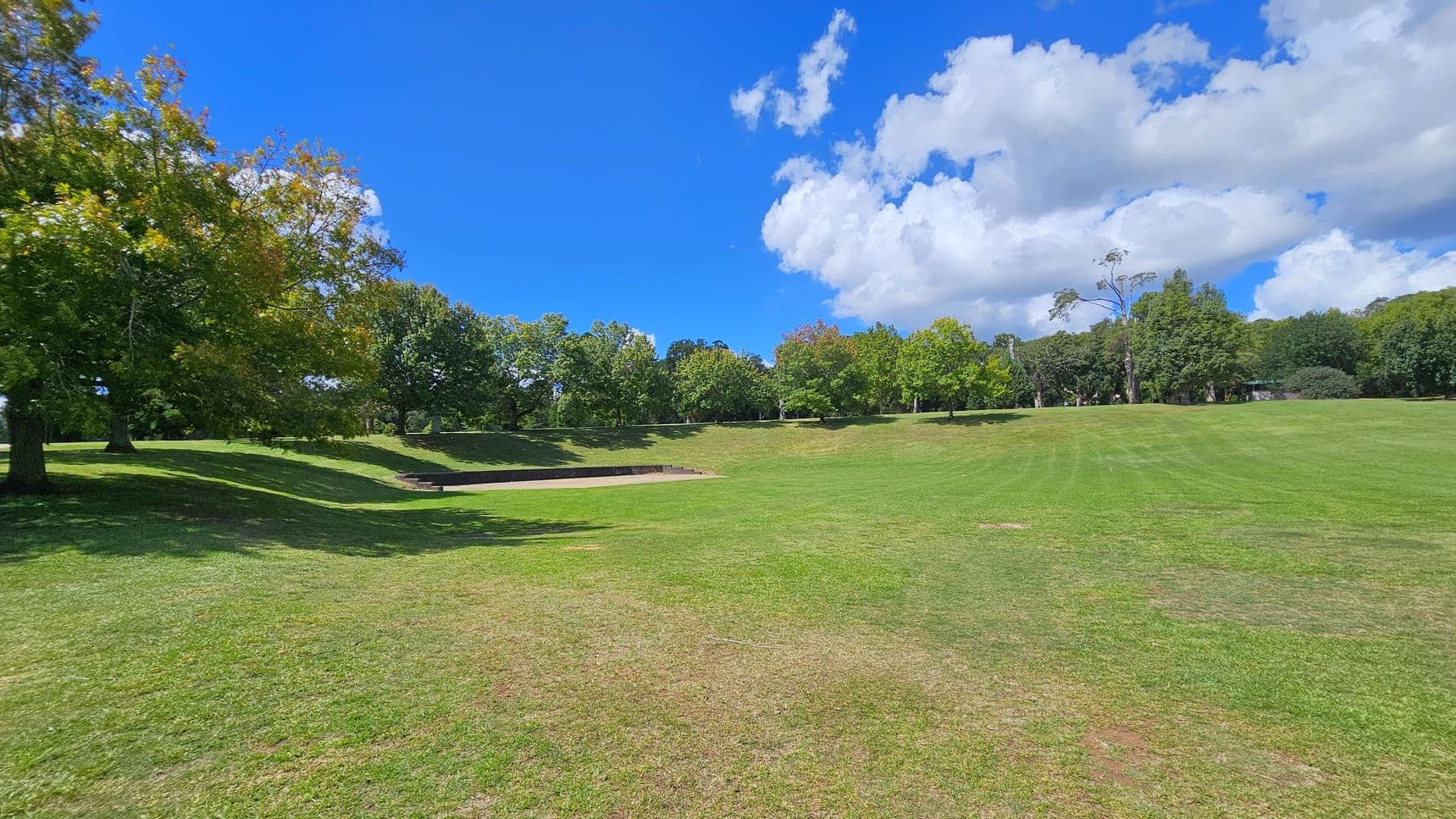 A grassy park landscape with a gentle slope. On the left, there are several large trees with green and yellow leaves, casting shadows on the ground. In the centre, a small, rectangular concrete structure is embedded in the grass. The right side features more open grassy space with a few scattered trees in the distance. The background is lined with a variety of trees, creating a natural border.